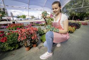 Young woman at a nursery holding flower plant in her hands as she kneels in the walkway between plants. photo