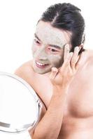 Beauty procedure and skin care. Close up portrait of happy laughing topless Asian man with face mask touching his face. Isolated on white background. photo