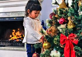 Asian little girl decorate the Christmas tree indoors. The morning before Xmas. Portrait loving girl close up. photo