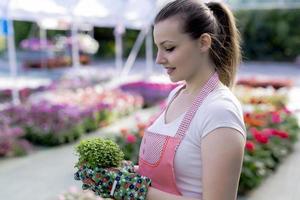 Young woman at a nursery holding flower plant in her hands as she kneels in the walkway between plants. photo