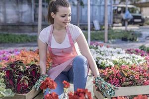 Young woman at a nursery holding flower plant in her hands as she kneels in the walkway between plants. photo