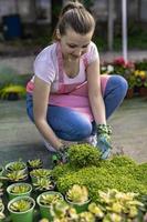 Young woman at a nursery holding flower plant in her hands as she kneels in the walkway between plants. photo