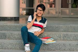 smiling young guy sits on a street with books and talking on the phone photo