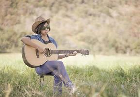 Women short hair wear hat and sunglasses sit playing guitar in grass field photo