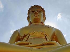 vista frontal estatua dorada del gran buda phra buddha dhammakaya thep mongkol en el templo wat pak nam phasi charoen. luz del sol cielo y fondo de nubes foto