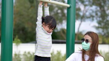 bambina asiatica appesa al bar dello sport che gioca su attrezzature per esercizi all'aperto, divertiti con sua madre al parco giochi video