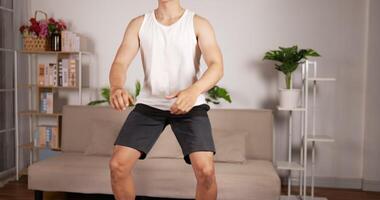 Young Asian man in earphone doing squat exercise in the living room at home. Fitness, sport, technology, and healthy lifestyle concept. video