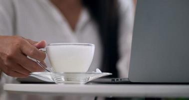 close-up de café, empresária bonita asiática bebendo café enquanto está sentado e trabalhando no laptop em seu local de trabalho. conceito de negócios e estilo de vida. video