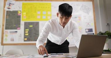 Porträt eines asiatischen Geschäftsmannes, der auf einen Laptop schaut und auf Papier schreibt, während er im Büroraum steht. Fokussierter Mann, der am Arbeitsplatz mit Computer-Notebook arbeitet. Mann überprüft Dokument. video