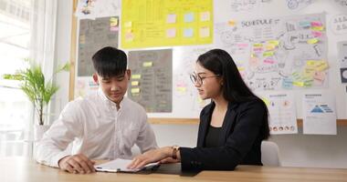 Asian businessman and businesswoman shaking hands to seal a deal with his partner while sitting at workplace desk in the office. Male and female sitting in room with sticky notes on board background. video