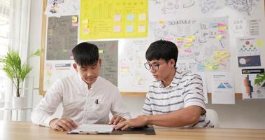 dois jovens asiáticos felizes apertando as mãos para fechar um acordo com seu parceiro enquanto estão sentados na mesa do local de trabalho no escritório. homem sentado na sala com notas autoadesivas no fundo da placa.