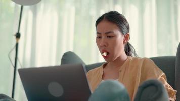 jovem mulher bonita sentada no sofá e comendo maçã enquanto digita no computador portátil, novo conceito normal e trabalhando em casa video