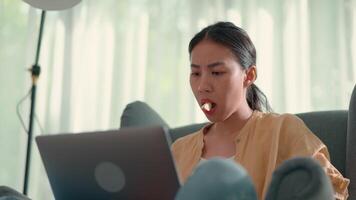 Young pretty woman sitting on sofa and eating apple while typing in laptop computer, new normal and working from home concept video