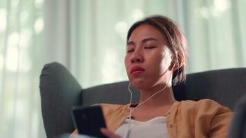 mujer joven sentada en un sofá en la sala de estar y usa auriculares para escuchar y hablar con alguien en el teléfono inteligente en casa video