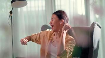 mujer joven sentada en un sofá en la sala de estar y usa auriculares para disfrutar bailando mientras escucha música desde la aplicación en el teléfono inteligente en casa