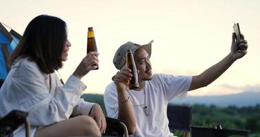 jeune bel homme et jolie femme s'amusant et buvant de la bière pendant un appel vidéo sur smartphone, ils rient avec plaisir ensemble devant la tente de camping