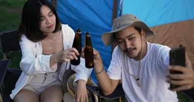 vue de dessus, jeune bel homme et jolie femme s'amusant et buvant de la bière pendant un appel vidéo sur smartphone, ils rient avec plaisir ensemble devant la tente de camping video