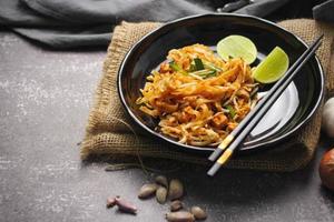 pad thai o pad thai al estilo tailandés servido con lima y condimentos en un plato negro, es una comida que se puede encontrar en la comida callejera de tailandia. foto