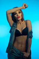 adorable young girl with red lips in black bra and jeans clothes posing on camera on blue background photo