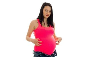 Cute brunette pregnant woman in bright pink shirt touching her belly isolated on white background photo