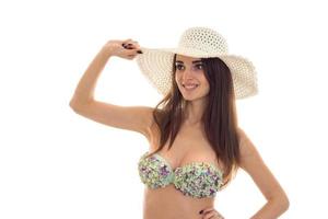 young cheerful brunette woman with big natural breasts in swimsuit with floral pattern and straw hat smiling and looking away isolated on white background photo