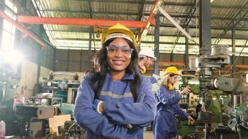 portrait d'une travailleuse noire professionnelle en uniforme de sécurité protecteur et casque regardant la caméra, les bras croisés et souriant, avec une équipe d'ingénieurs derrière elle dans une usine de fabrication de métaux. video