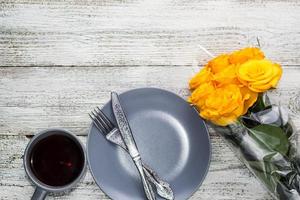 a plate with a knife and fork and tea with a bouquet of yellow roses on a white wooden background