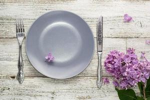 spring table setting with plate fork and knife decorated with lilac flowers on white wooden background