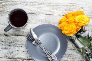 a plate with a knife and fork and tea with a bouquet of yellow roses on a white wooden background photo