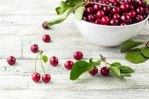 tazón blanco de cerezas dulces frescas con hojas en gotas de agua sobre fondo de madera foto