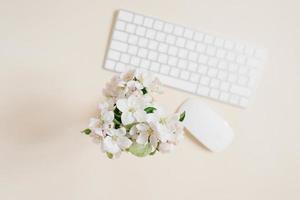 teclado y ratón y flores blancas de manzana en un jarrón fuera de foco sobre una mesa beige. diseño y concepto de diseño. endecha plana foto