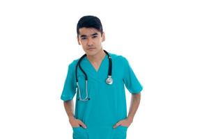 a young doctor in the blue dress with stethoscope keeps his hands in his pockets and looking directly photo