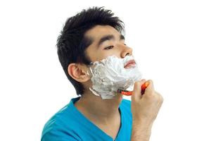 un joven guapo con el pelo negro se para de lado con espuma en la cara y se afeita la barba foto