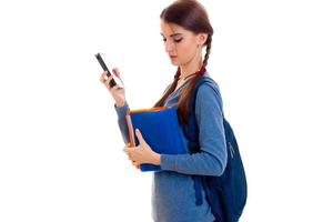 young smart brunette student girl with backpack on her shoulders reads a book and talking phone isolated on white background photo