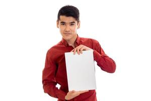 Joven y elegante modelo masculino moreno con camisa roja posando con una pancarta vacía en las manos y mirando a la cámara aislada de fondo blanco foto