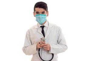 retrato de un joven y alegre doctor moreno con uniforme blanco y máscara azul con estetoscopio en el cuello mira la cámara aislada en un fondo vacío foto