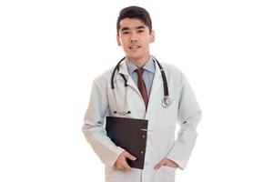 portrait of serious young brunette male doctor in uniform with stethoscope posing isolated on white background photo