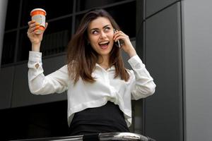 mujer de negocios feliz en una camisa sosteniendo una taza de café y hablando por teléfono