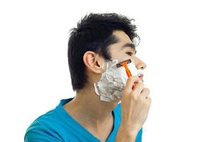 un joven con el pelo negro se para de lado frente a la cámara con la espuma en las mejillas y se afeita la barba de cerca foto