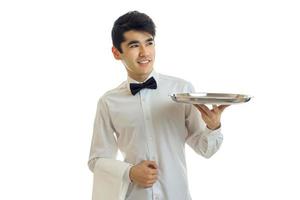 joven camarero moreno alegre con uniforme y bandeja en las manos sonriendo y mirando a un lado foto