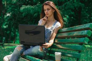 linda jovencita sentada en un banco en un parque con una laptop negra foto