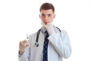 the handsome young doctor with a mask on his face and wearing a robe looks straight and holding a syringe photo