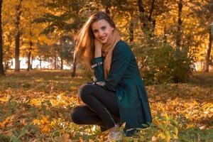una joven linda con un manto negro se sienta en el parque en otoño en el césped y sonríe foto