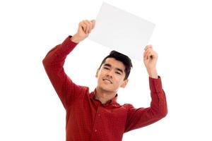 Joven modelo masculino moreno feliz con camisa roja posando con una pancarta vacía en las manos y mirando y sonriendo a la cámara aislada de fondo blanco foto