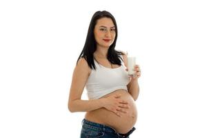 young pregnant girl with dark hair touching her big belly drinks milks and looking at the camera isolated on white background photo