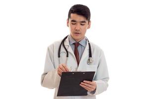 retrato de un joven y apuesto doctor moreno con uniforme blanco posando en la cámara con estetoscopio en el cuello y haciendo notas aisladas en un fondo vacío foto