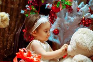 Young girl playing with her teddy bear under white Christmas tree photo