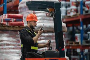 Manager or engineer man employee use walkie-talkie to give order to co-worker and checking stock on forklift. Worker wearing high visibility clothing, helmet count up goods or boxes for delivery. photo