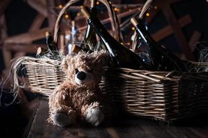 Teddy bear, champagne bottle with Christmas basket, gold garlands on dark brown wooden background, new year celebration concept photo