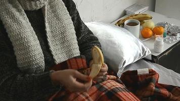 mujer joven enferma en el suéter se acuesta en la cama y come un plátano en casa video